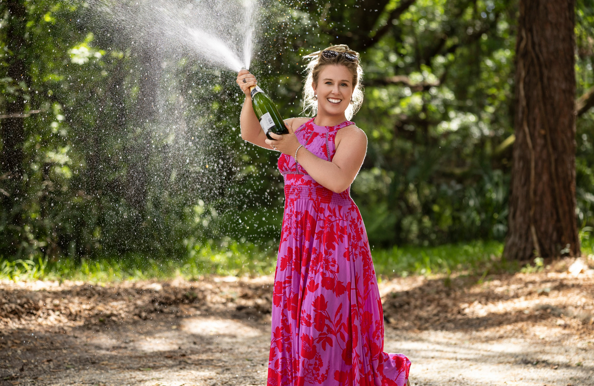 Heidi spraying champagne in a pink floral maxi dress with sunlight glittering off the spray — celebratory realtor branding portrait, Beaufort SC