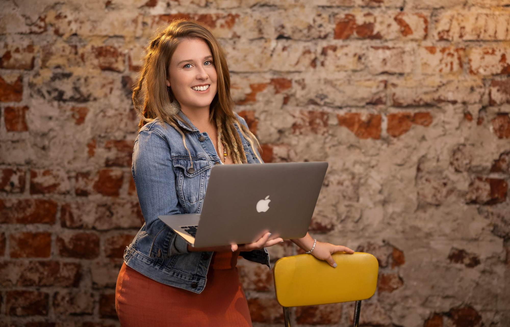 Heidi with a laptop in a denim jacket against an exposed brick wall — realtor branding session, Beaufort SC