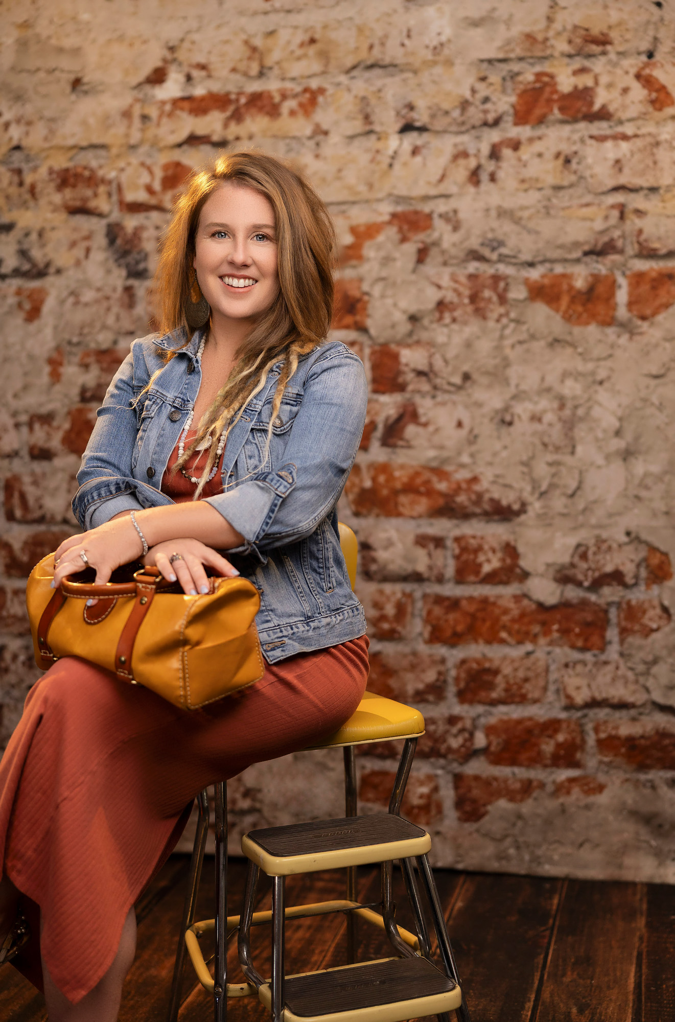 Heidi seated on a vintage yellow kitchen step stool against an exposed brick wall — personal-prop realtor branding portrait, Beaufort SC