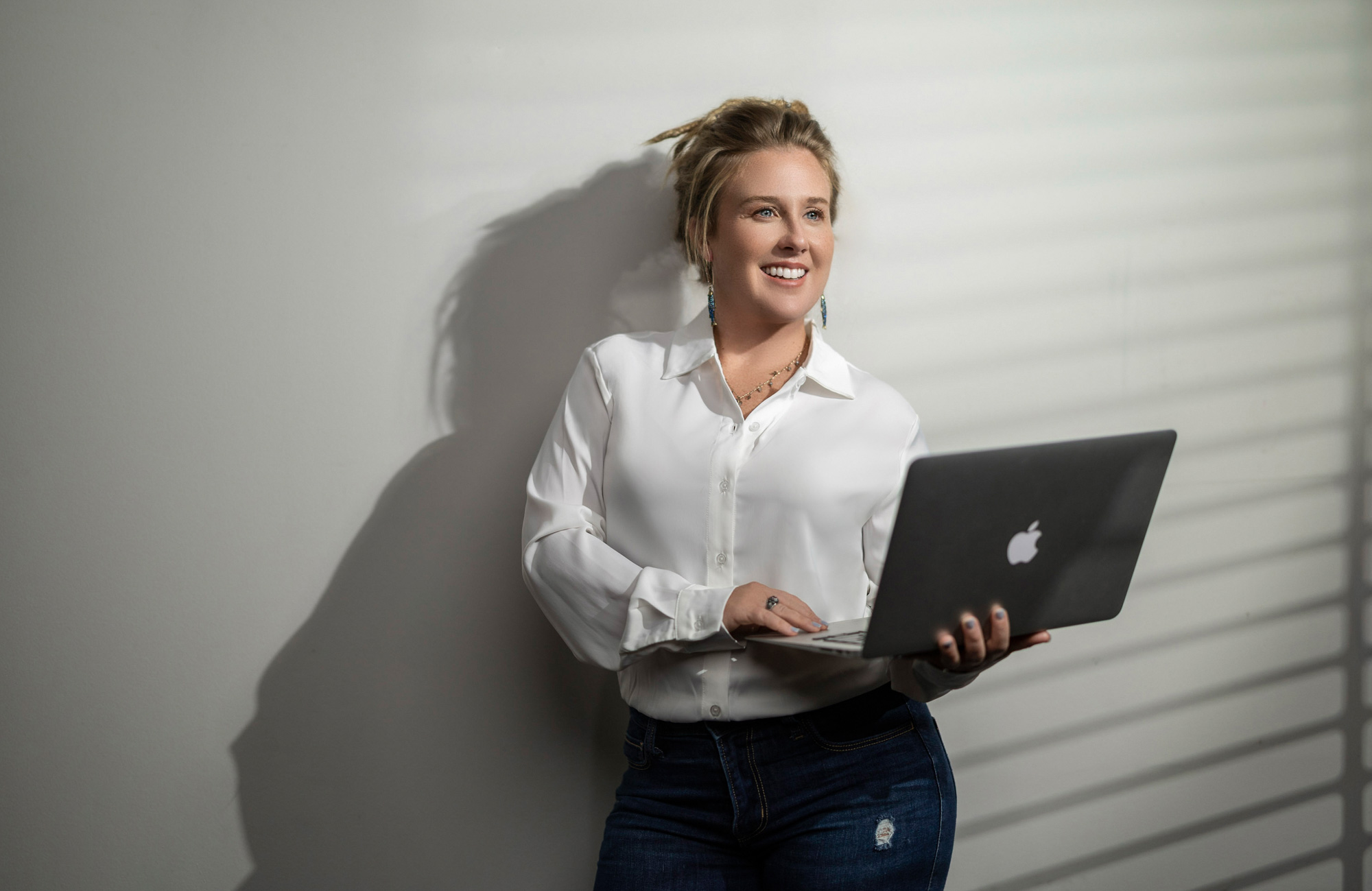Heidi in a white shirt and jeans holding a laptop, light wall background — realtor branding portrait, Beaufort SC