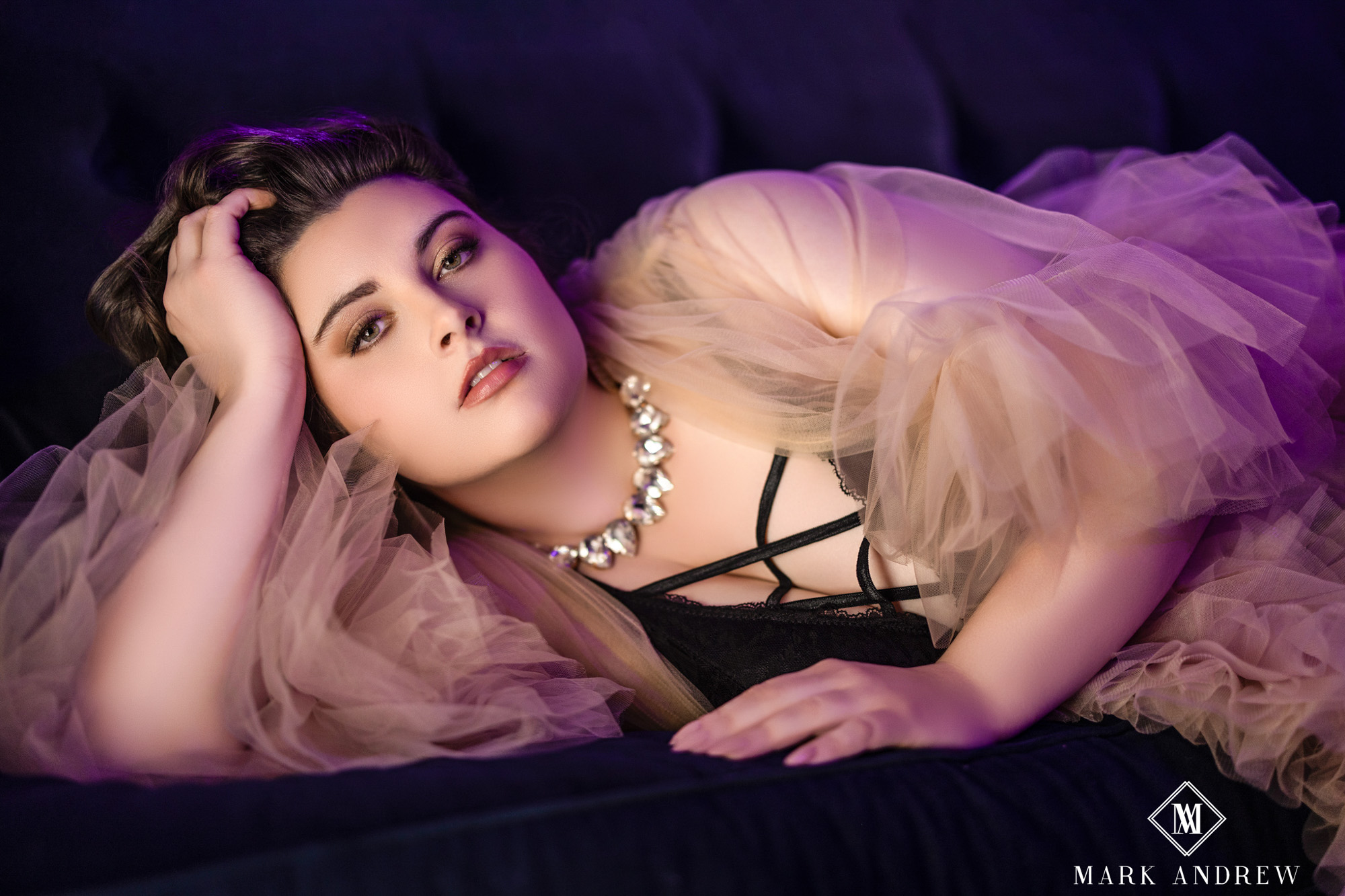 Katherine reclining on a navy velvet sofa in peach tulle and a diamond choker, lit in moody purple — cinematic boudoir portrait