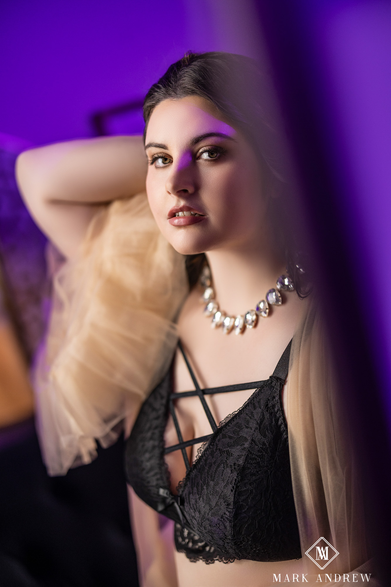 Katherine in a black lace bralette with a diamond choker and tulle drape, photographed in dramatic purple light — mid-session boudoir portrait