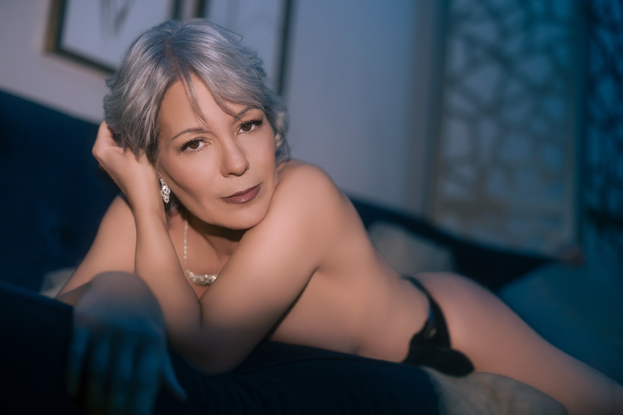 Mature boudoir client with silver hair, pearl necklace, on velvet sofa in cool blue light